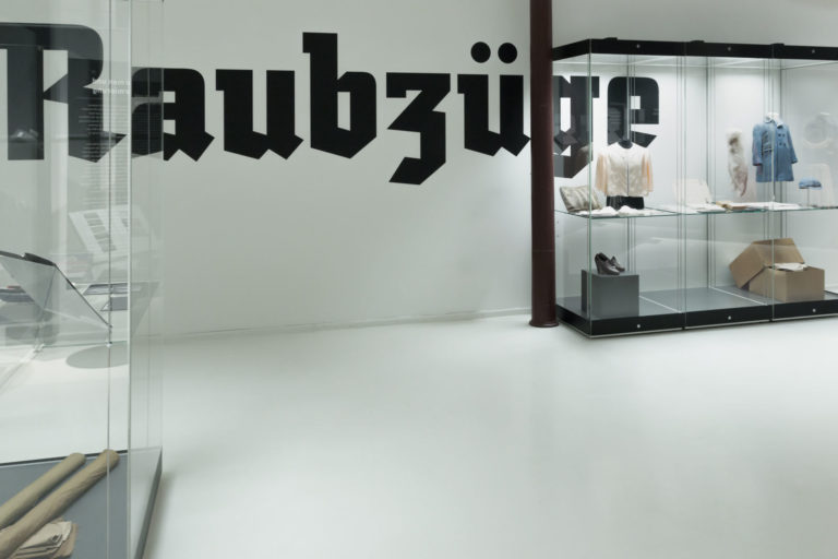 Das Referenzbild für Ausstellungsgestaltung aus der Sonderausstellung „Glanz und Grauen. Mode im Dritten Reich“ zeigt Großraumvitrinen und eine Wandbeschriftung.