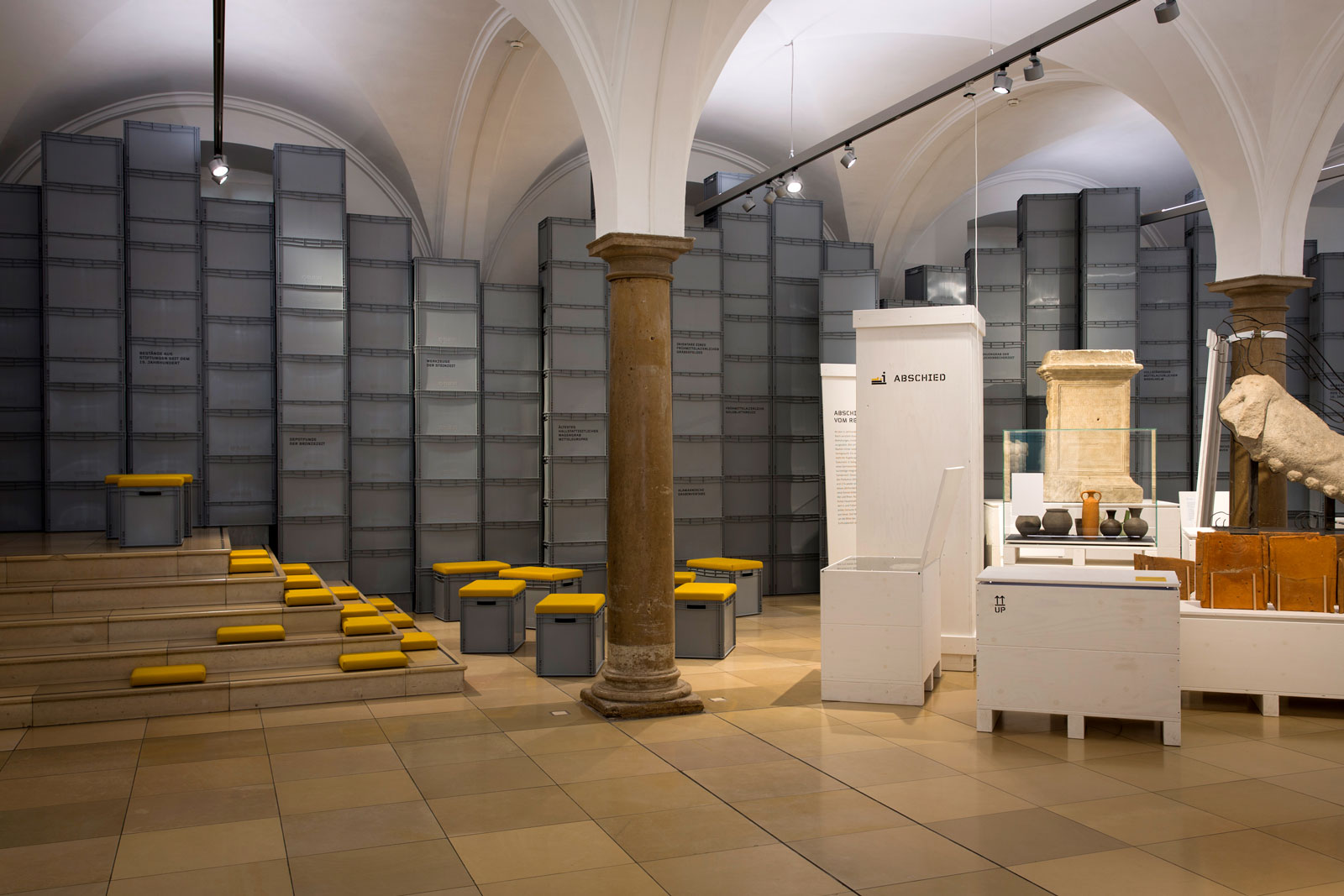 Das Referenzbild für Ausstellungsgestaltung aus der Interimsausstellung „Römerlager – Das Römische Augsburg in Kisten“ zeigt eine Rauminszenierung mit Archivboxen und Lichtplanung.