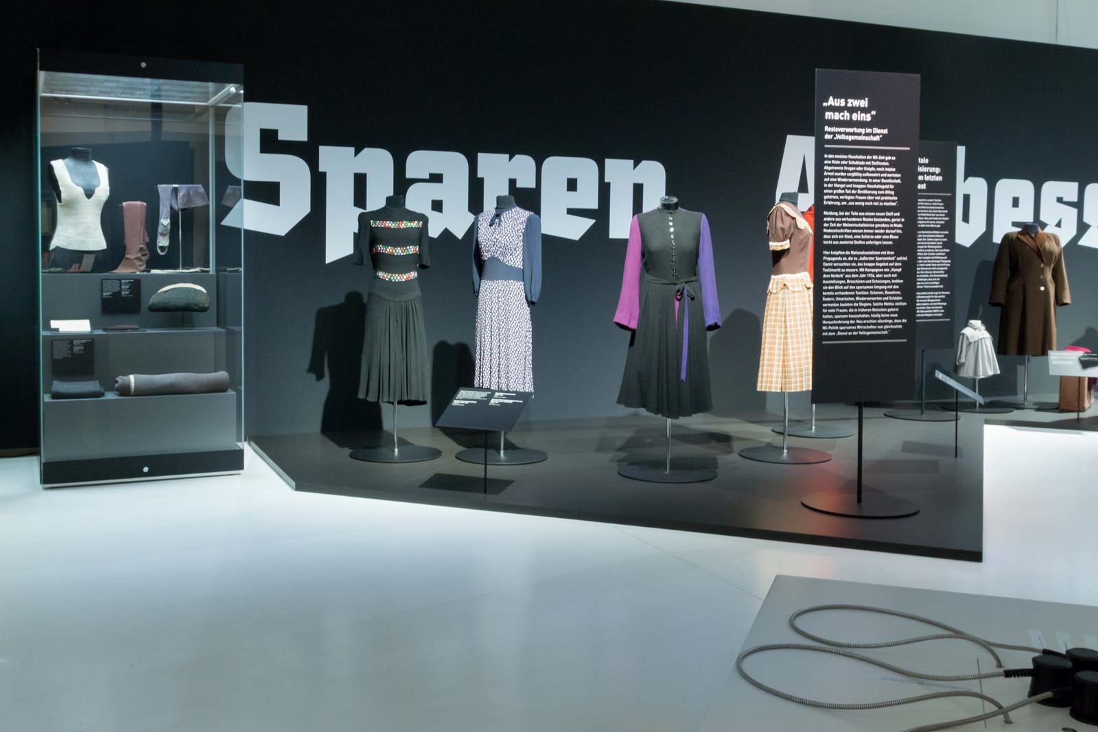 Das Referenzbild für Ausstellungsgestaltung aus der Sonderausstellung „Glanz und Grauen. Mode im Dritten Reich“ zeigt historische Kleidungsstücke in Ganzglasvitrinen und Vitrinen.
